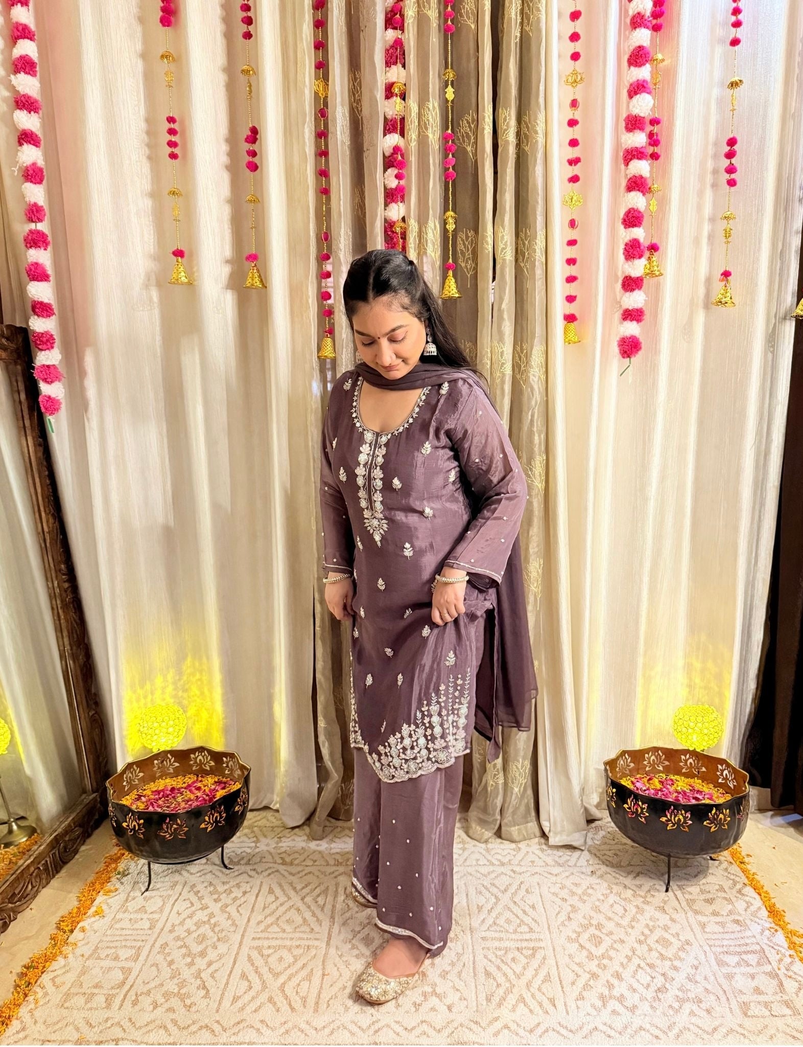 Woman in a purple traditional outfit standing in front of decorative curtains with floral arrangements. Blooming Mauve Embroidered Chinon Suit