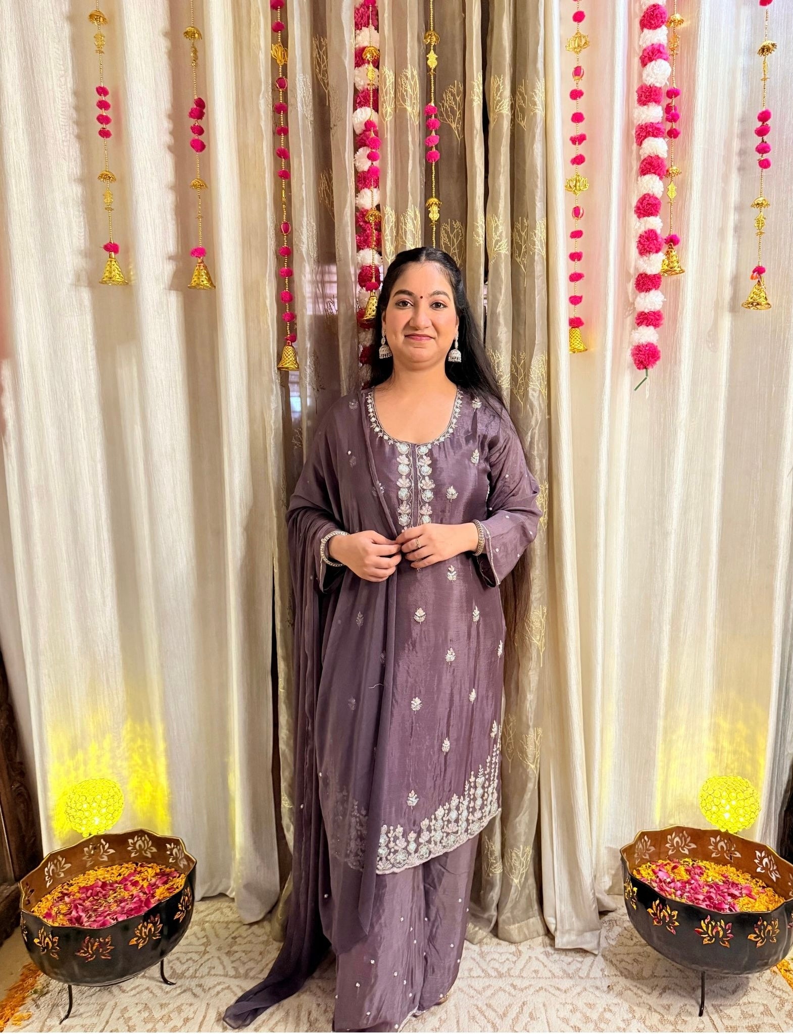 Woman in a traditional outfit standing in front of decorative curtains with floral arrangements. Blooming Mauve Embroidered Chinon Suit