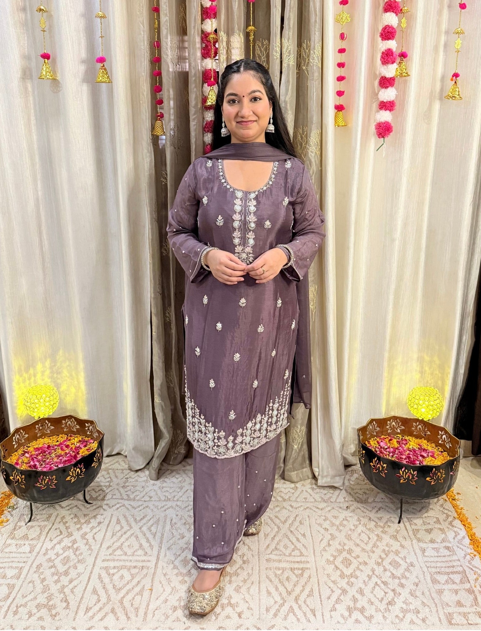 Woman in a traditional outfit standing in front of decorative curtains with floral arrangements. Blooming Mauve Embroidered Chinon Suit