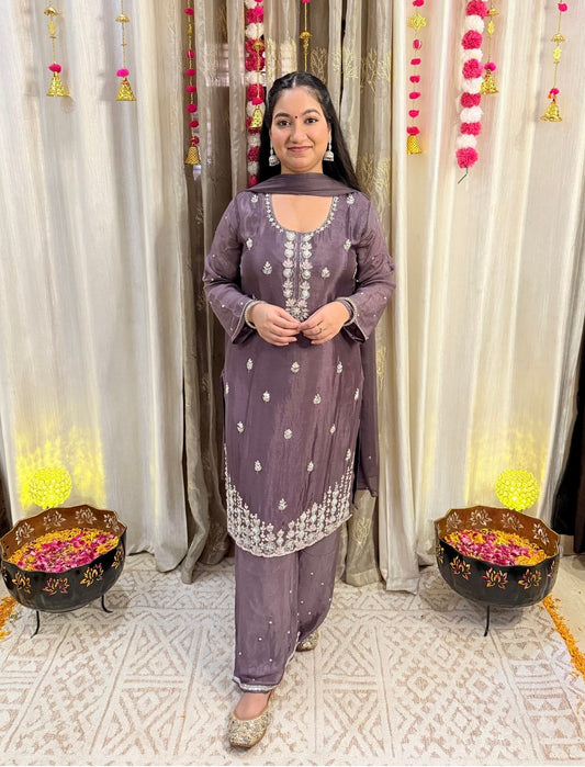 Woman in a traditional outfit standing in front of decorative curtains with floral arrangements. Blooming Mauve Embroidered Chinon Suit