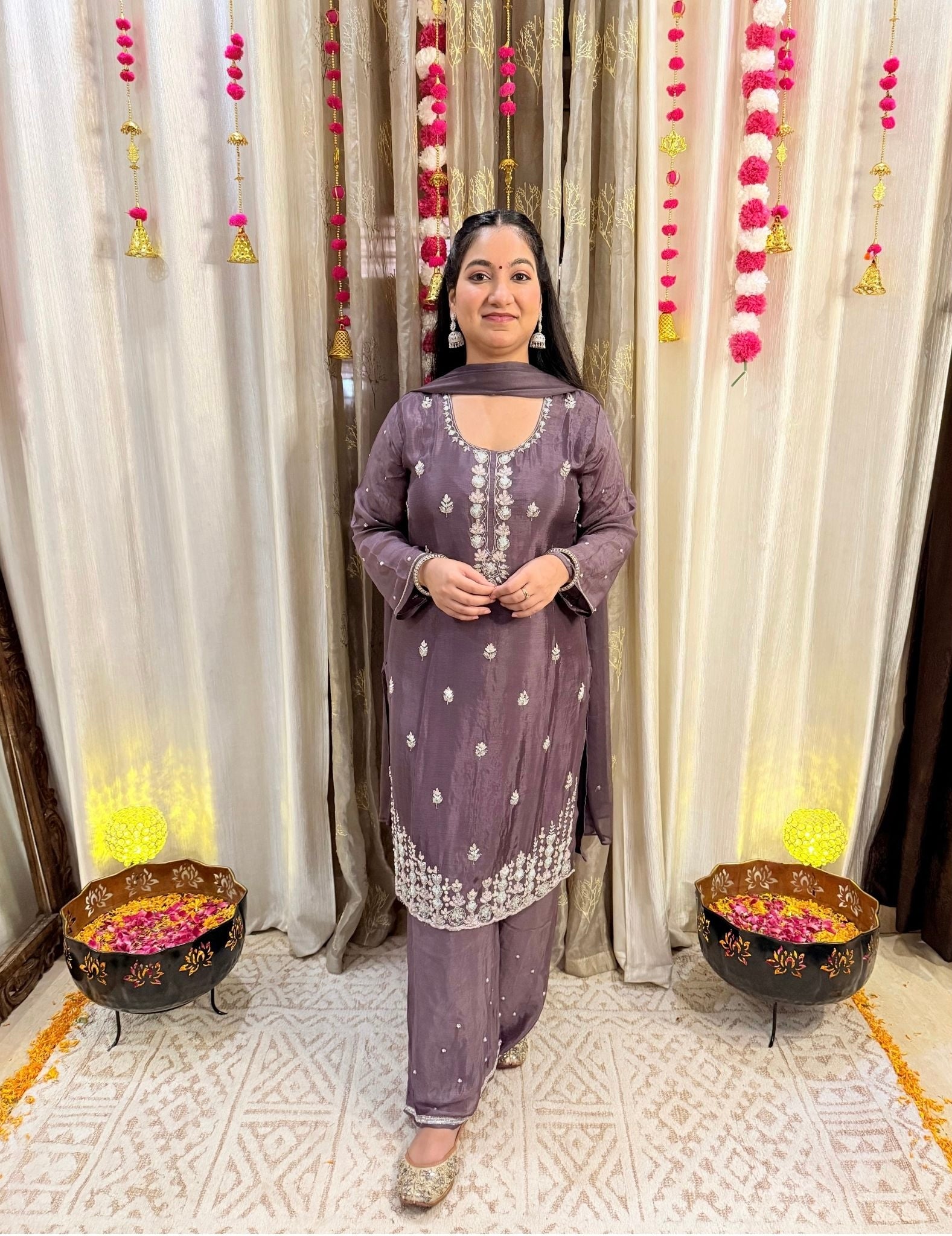Woman in a purple traditional outfit standing in front of decorated curtains with floral arrangements. Blooming Mauve Embroidered Chinon Suit