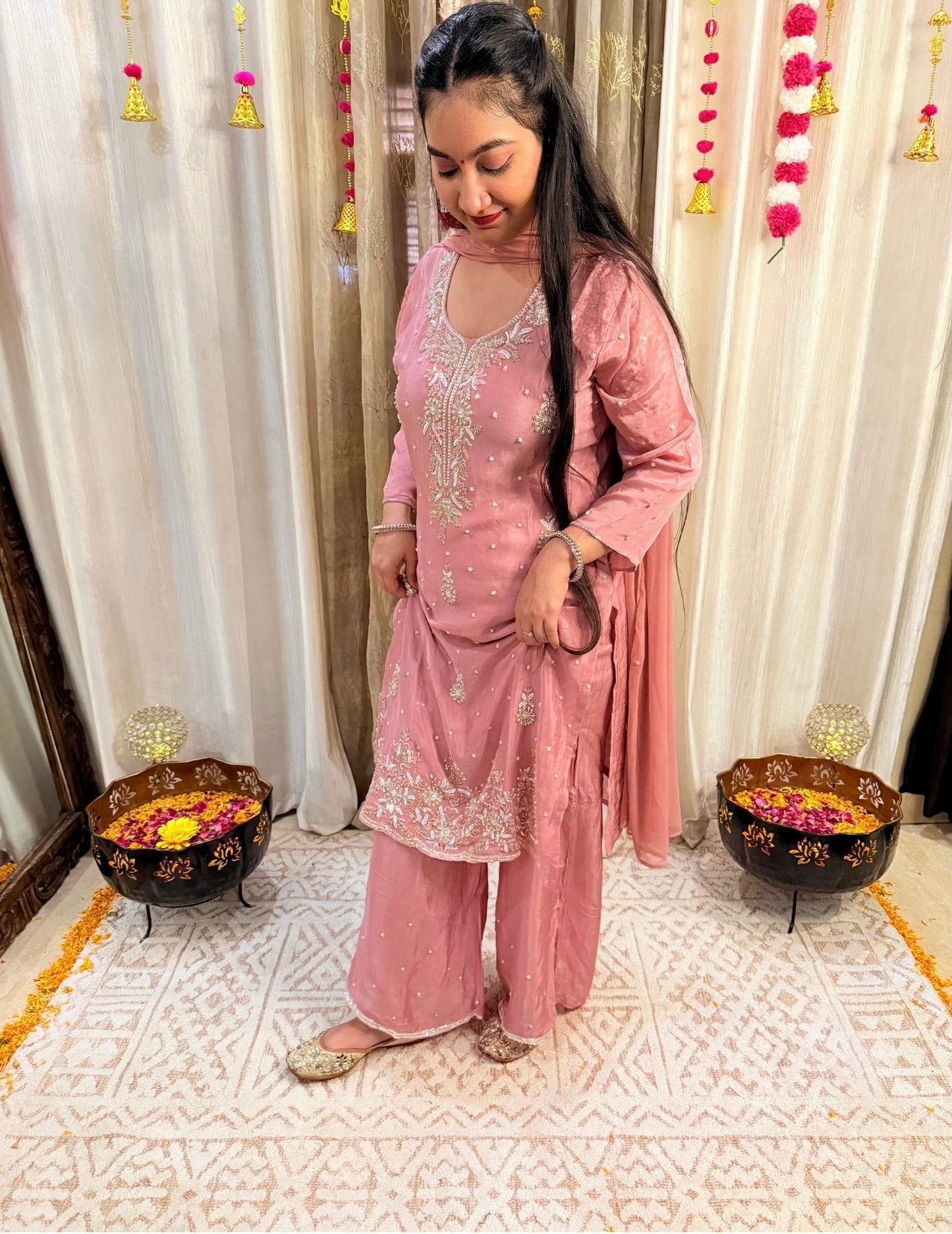 Woman in a pink traditional outfit standing in a decorated room with white curtains and floral decorations. DUSTY_ROSE_EMBROIDERED_CHINON_SUIT