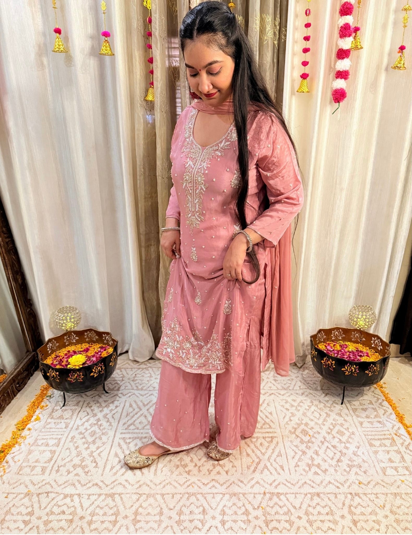Woman in a pink traditional outfit standing in a decorated room with white curtains and floral decorations. DUSTY_ROSE_EMBROIDERED_CHINON_SUIT