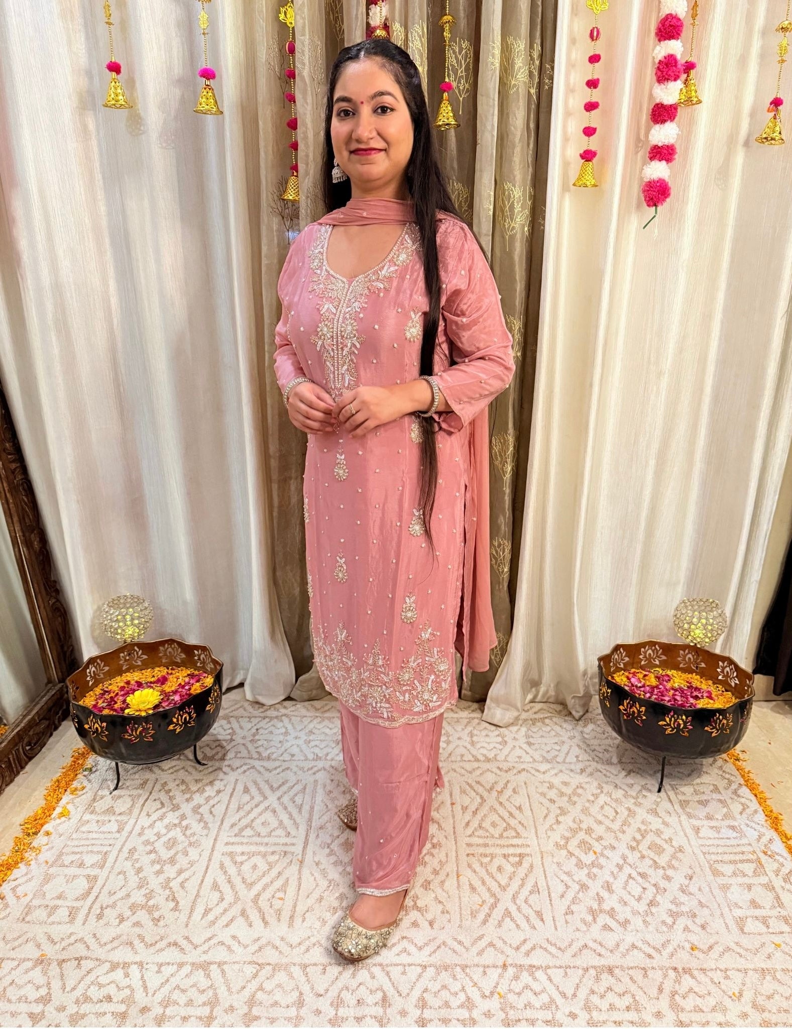 Woman in a pink traditional outfit standing in front of decorative curtains with floral arrangements on a table. DUSTY_ROSE_EMBROIDERED_CHINON_SUIT