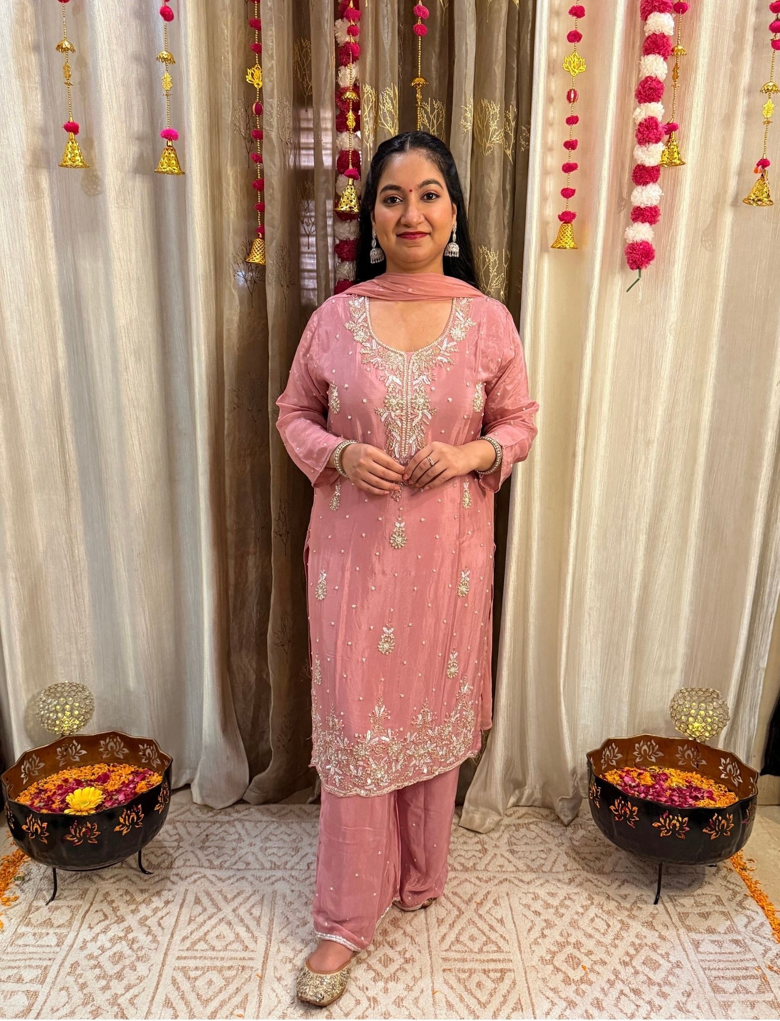 Woman in a pink traditional outfit standing in front of decorative curtains with floral arrangements.DUSTY_ROSE_EMBROIDERED_CHINON_SUIT