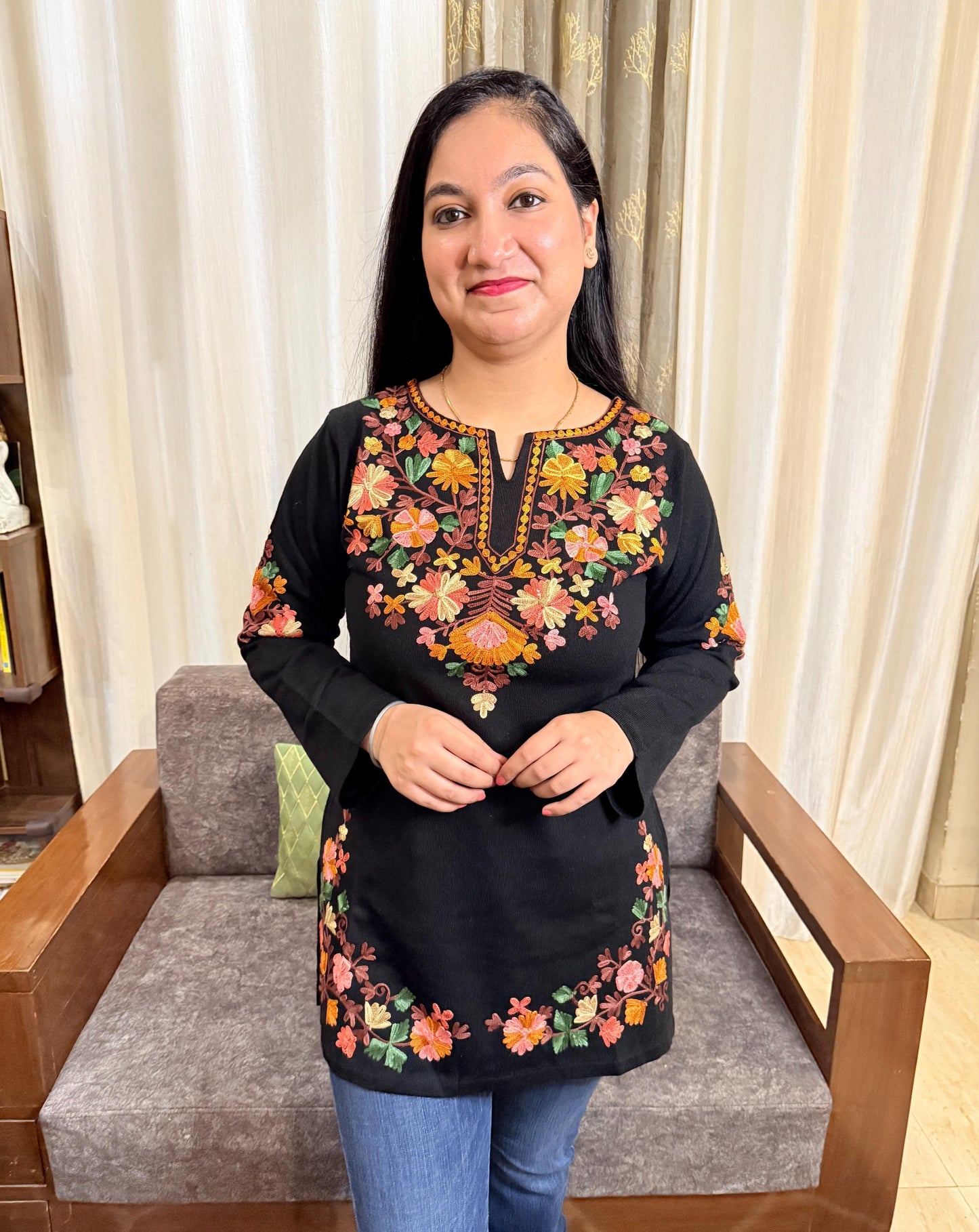 Woman wearing a black top with colorful floral embroidery sitting on a gray sofa. GulmoharBlackEmbroideredKashmiriWoollenKurti1