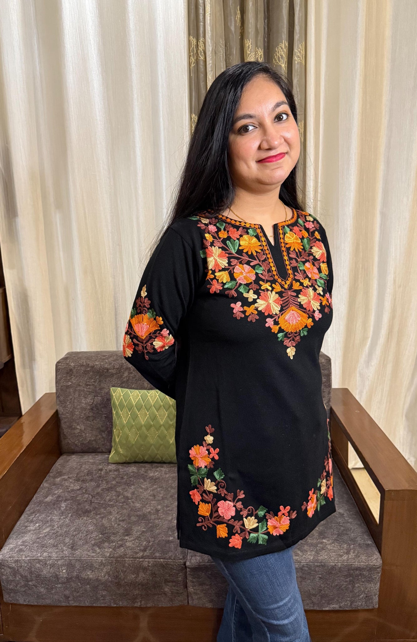 Woman wearing a black embroidered top sitting on a gray sofa with a curtain background GulmoharBlackEmbroideredKashmiriWoollenKurti3