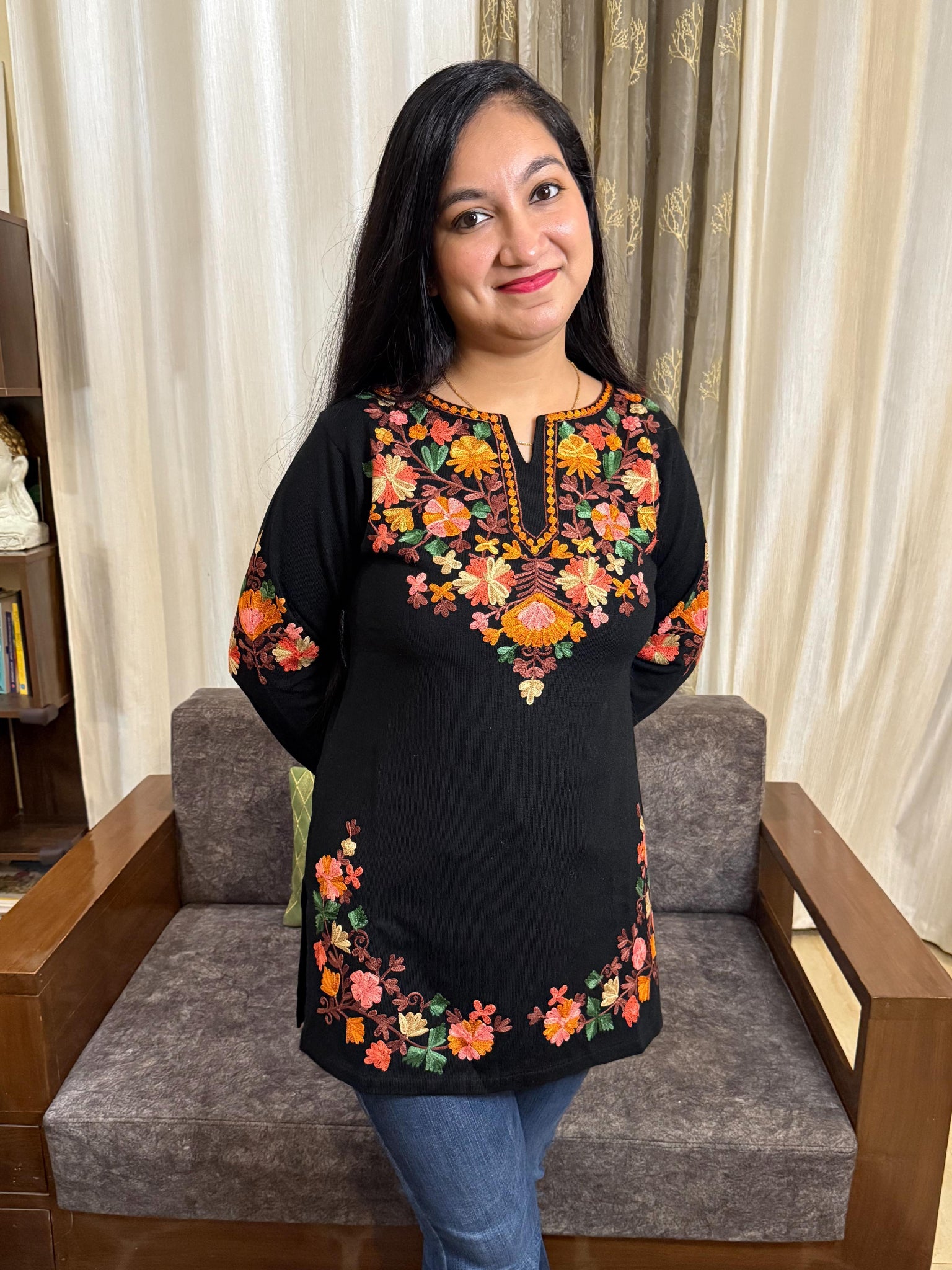 Woman wearing a black top with colorful floral embroidery in a living room setting. GulmoharBlackEmbroideredKashmiriWoollenKurti3