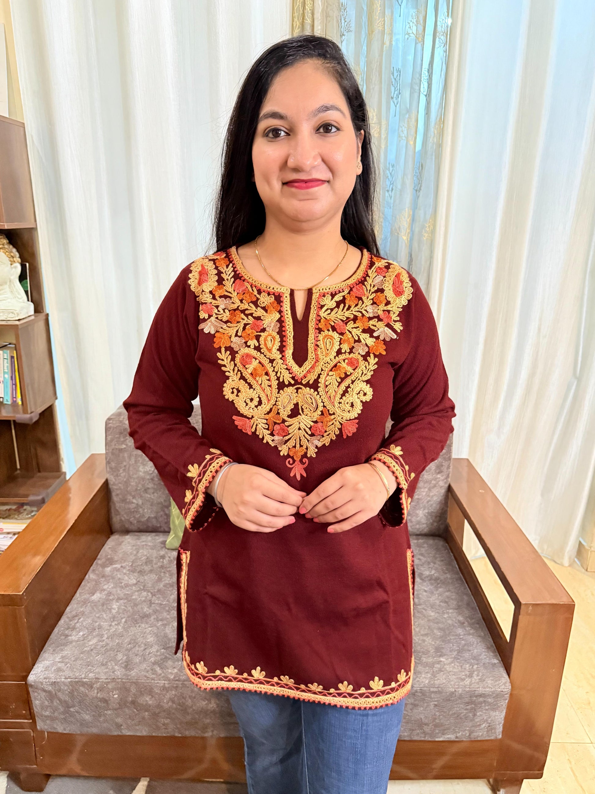 Woman wearing a maroon top with intricate gold embroidery in a room. GulmoharMaroonEmbroideredKashmiriWoollenKurti