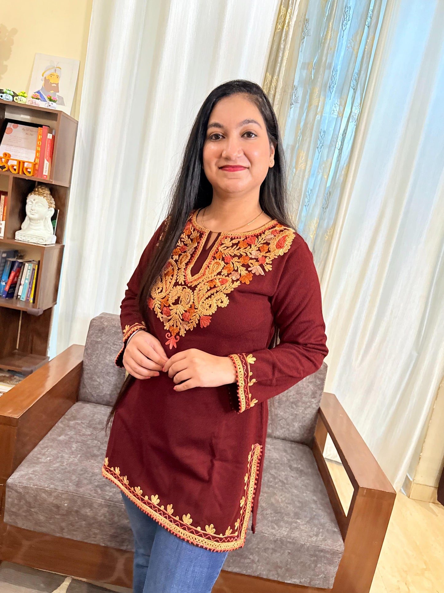 Woman wearing a maroon top with gold embroidery sitting on a chair indoors. GulmoharMaroonEmbroideredKashmiriWoollenKurti1