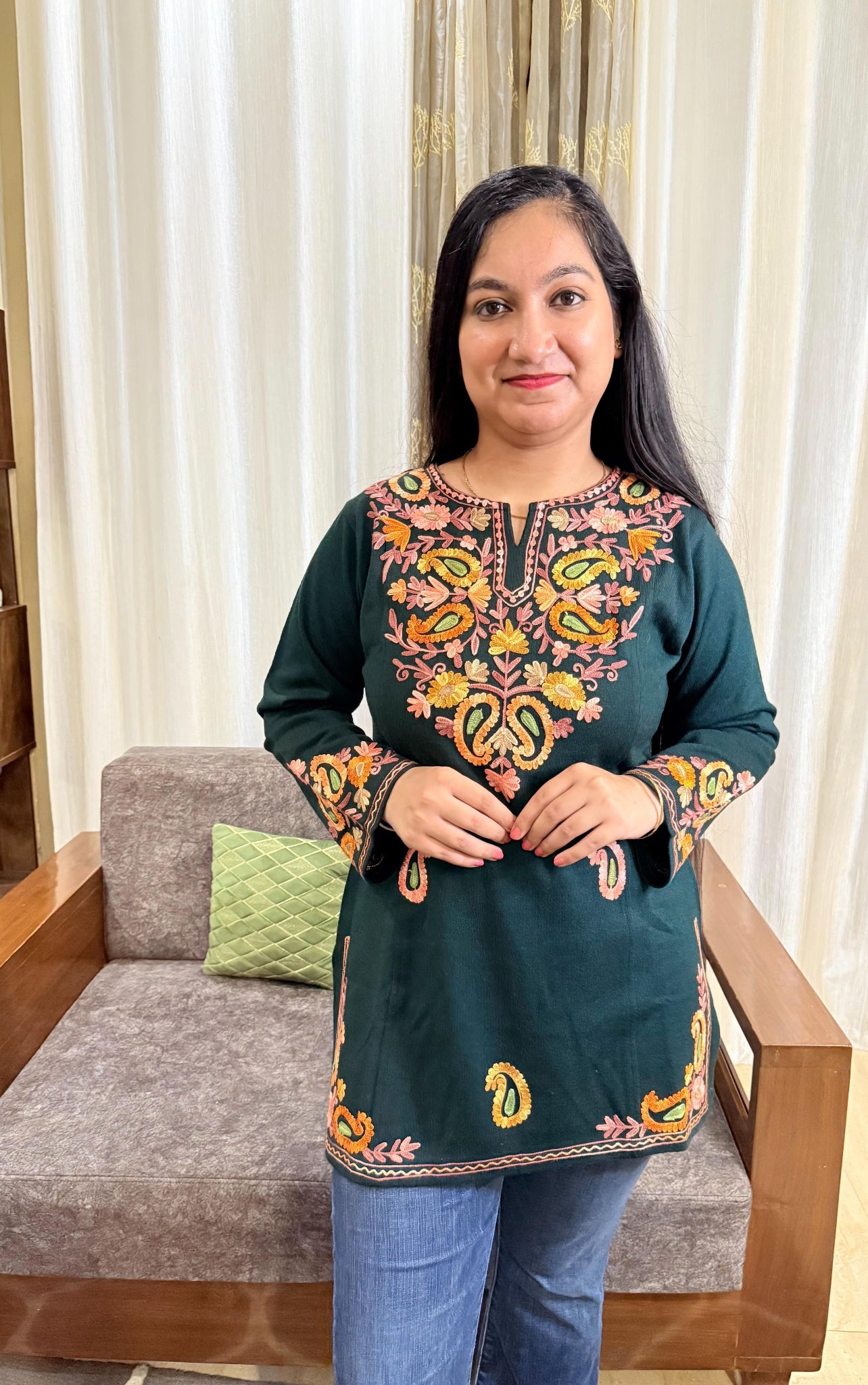 Woman wearing a green embroidered top standing in a room with curtains and a chair. GulmoharTealEmbroideredKashmiriWoollenKurti3