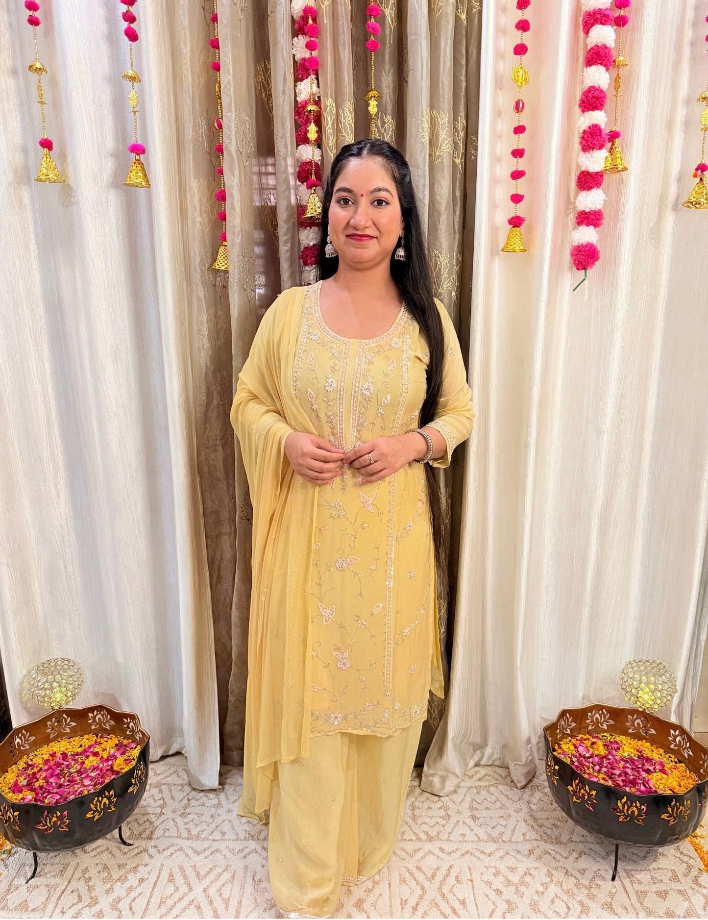 Woman in a yellow traditional outfit standing in front of decorated curtains with floral arrangements on either side. Lemonlight_Embroidered_Chinon_Suit