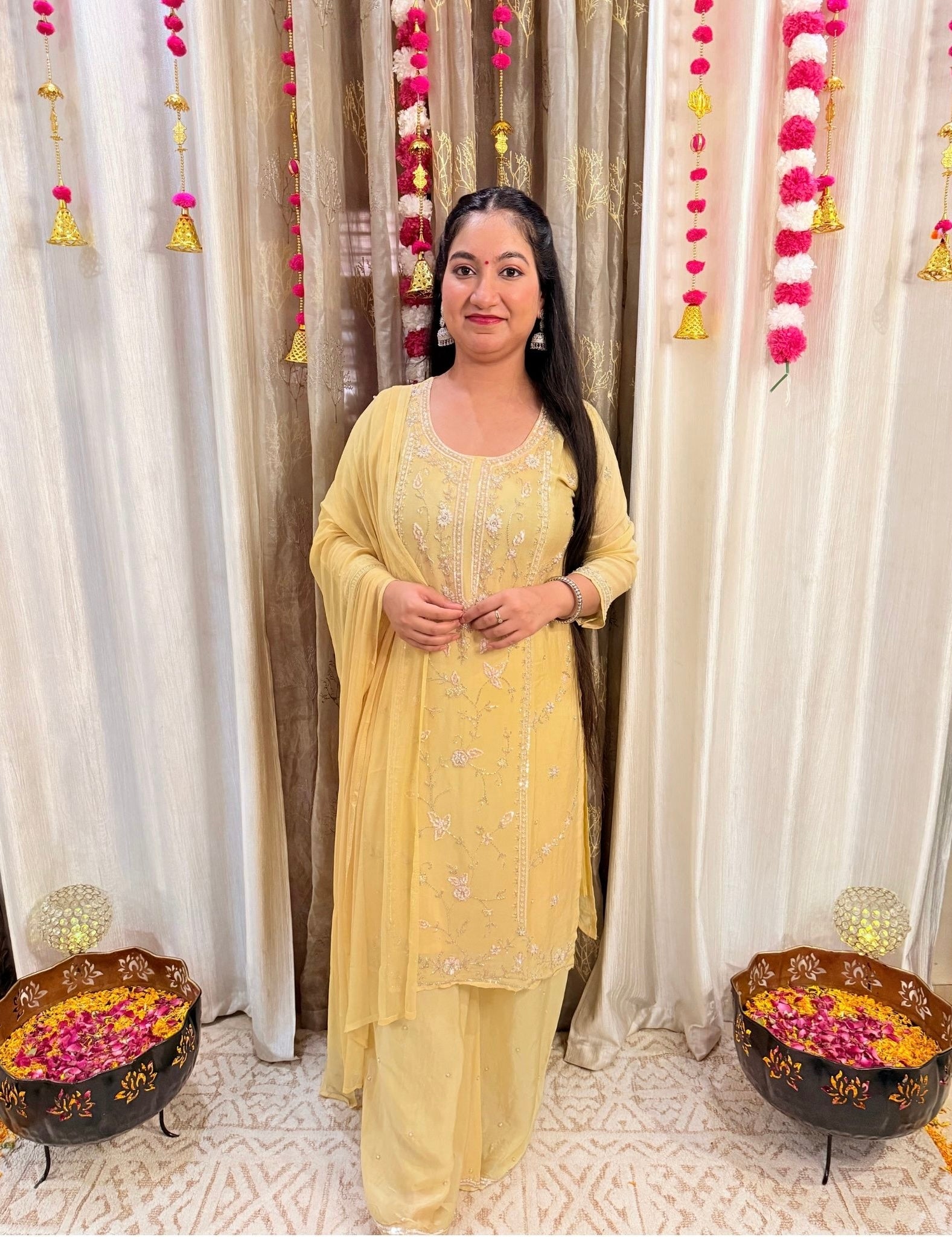 Woman in a yellow traditional outfit standing in front of decorated curtains with floral arrangements on either side. Lemonlight_Embroidered_Chinon_Suit