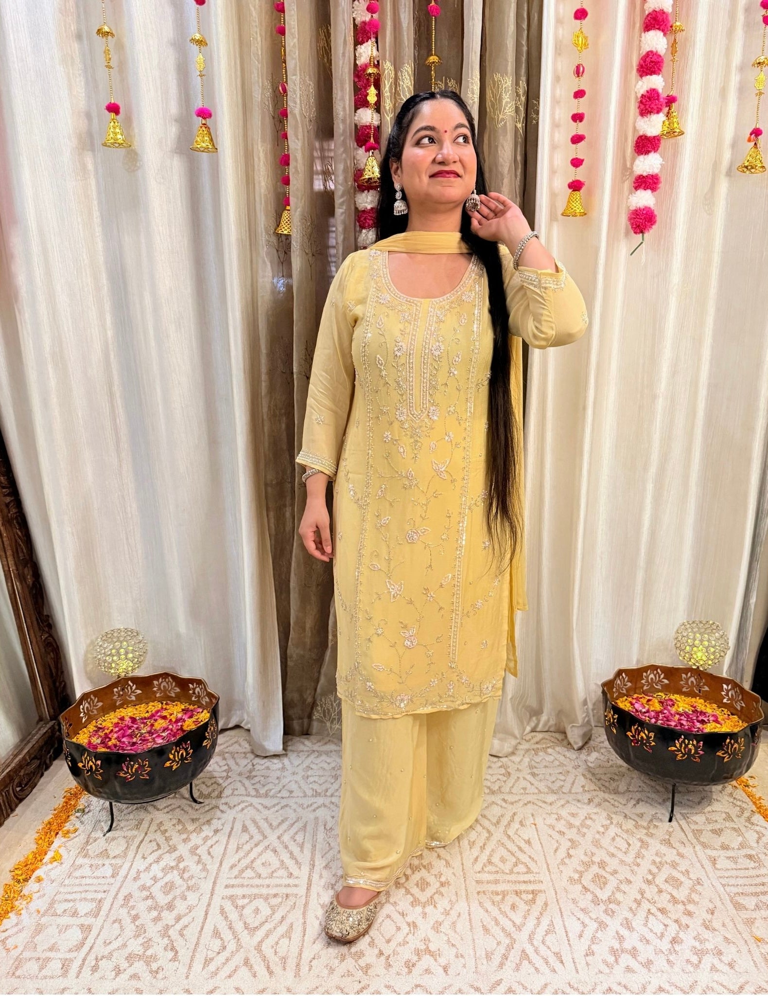 Woman in a yellow traditional outfit standing in front of decorative curtains with floral arrangements. Lemonlight_Embroidered_Chinon_Suit