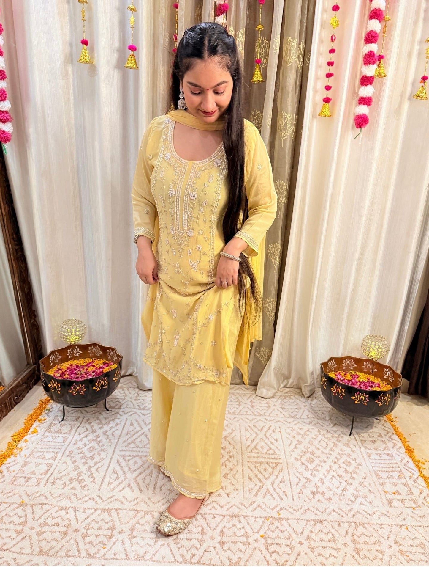 Woman in a yellow traditional outfit standing in a decorated room with curtains and floral decorations. Lemonlight_Embroidered_Chinon_Suit