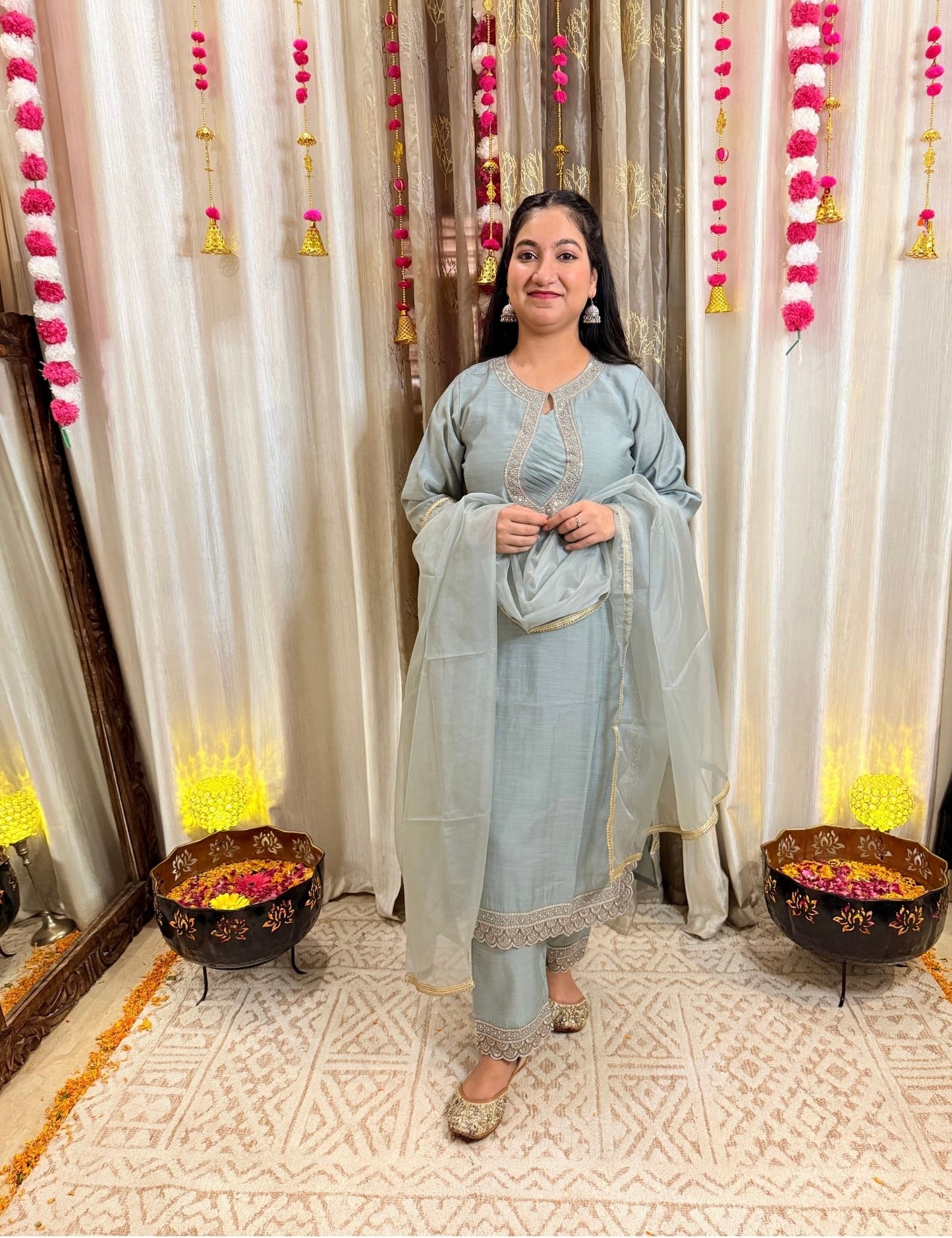 Woman in a light blue traditional outfit standing in front of decorative curtains with pink and yellow decorations. MEGHGREYISHBLUEFESTIVESUIT