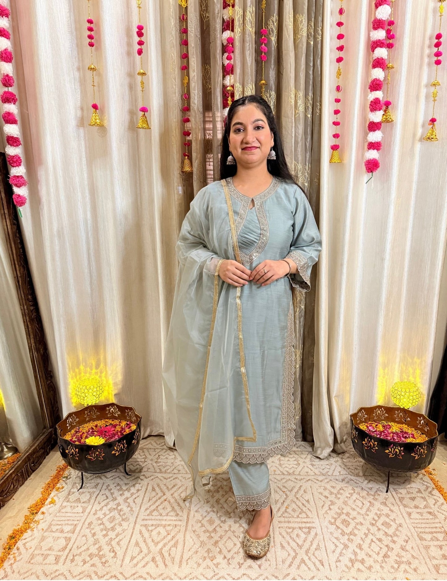 Woman in a light blue traditional outfit standing in front of decorative curtains with floral arrangements.MEGHGREYISHBLUEFESTIVESUIT