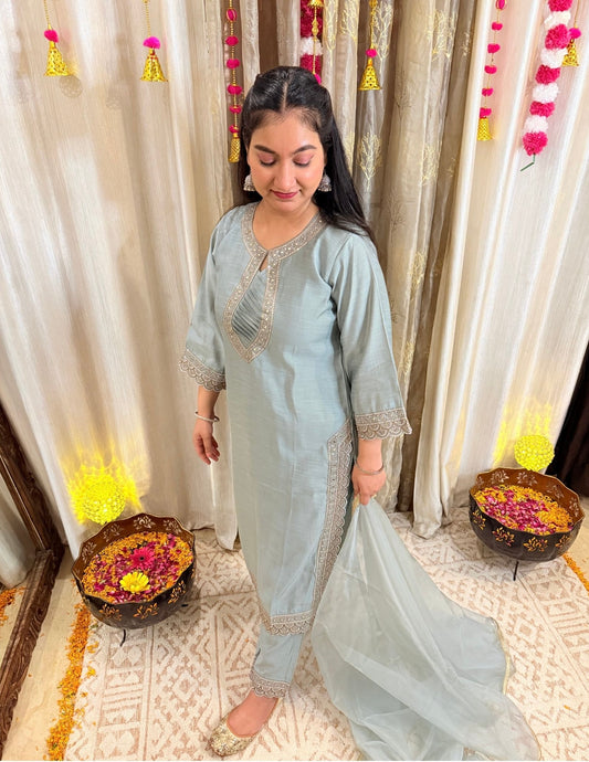 Woman in a light blue traditional outfit standing in front of decorative curtains. MEGHGREYISHBLUEFESTIVESUIT