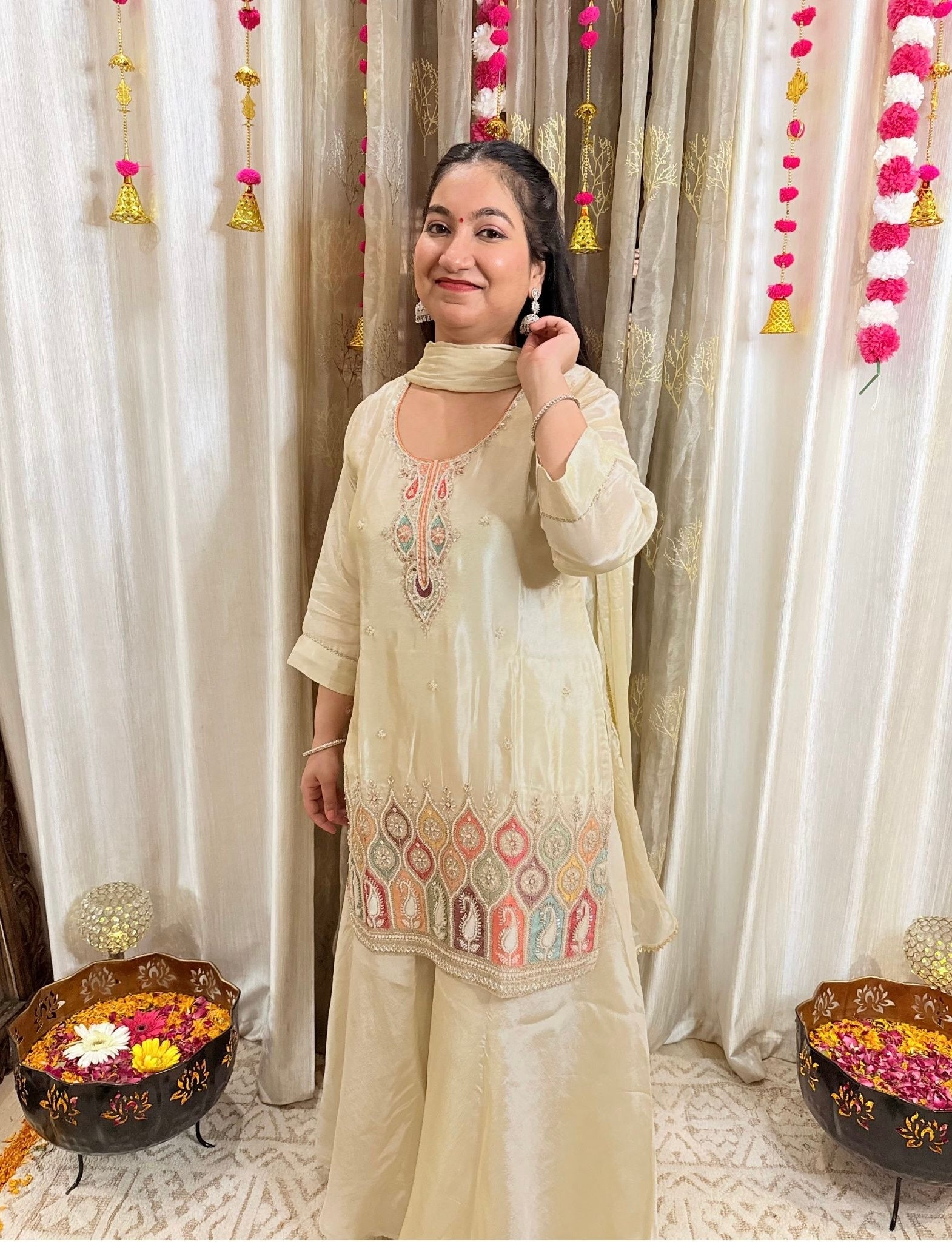 Woman in a cream traditional outfit with intricate patterns standing in front of decorative curtains.