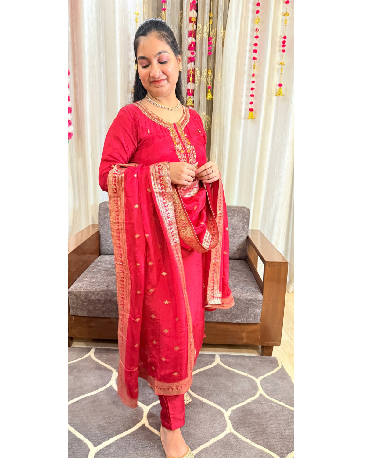 Woman in a red traditional outfit with a dupatta, standing in a room with curtains and a sofa. ZYRAFESTIVESUITSETTHEMKMAAN
