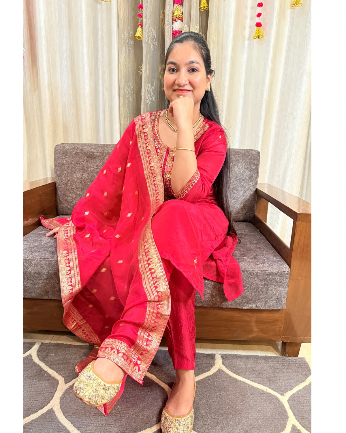 Woman in a pink traditional outfit sitting on a gray sofa with white curtains in the background. ZYRAFESTIVESUITSETTHEMKMAAN