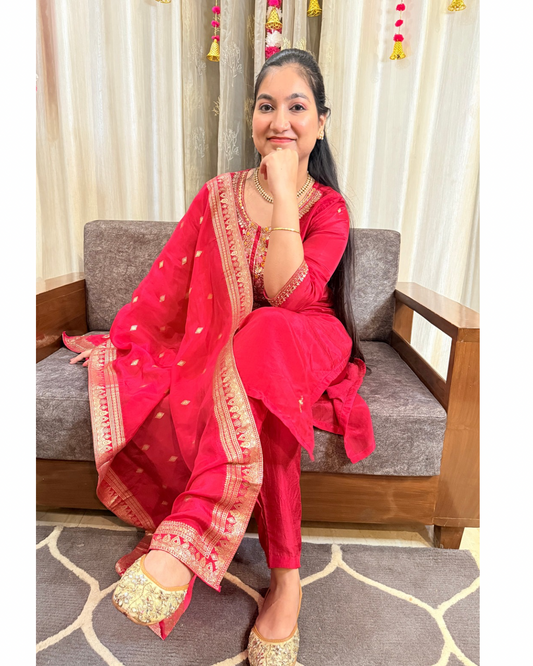 Woman in a pink traditional outfit sitting on a gray sofa with white curtains in the background. ZYRAFESTIVESUITSETTHEMKMAAN