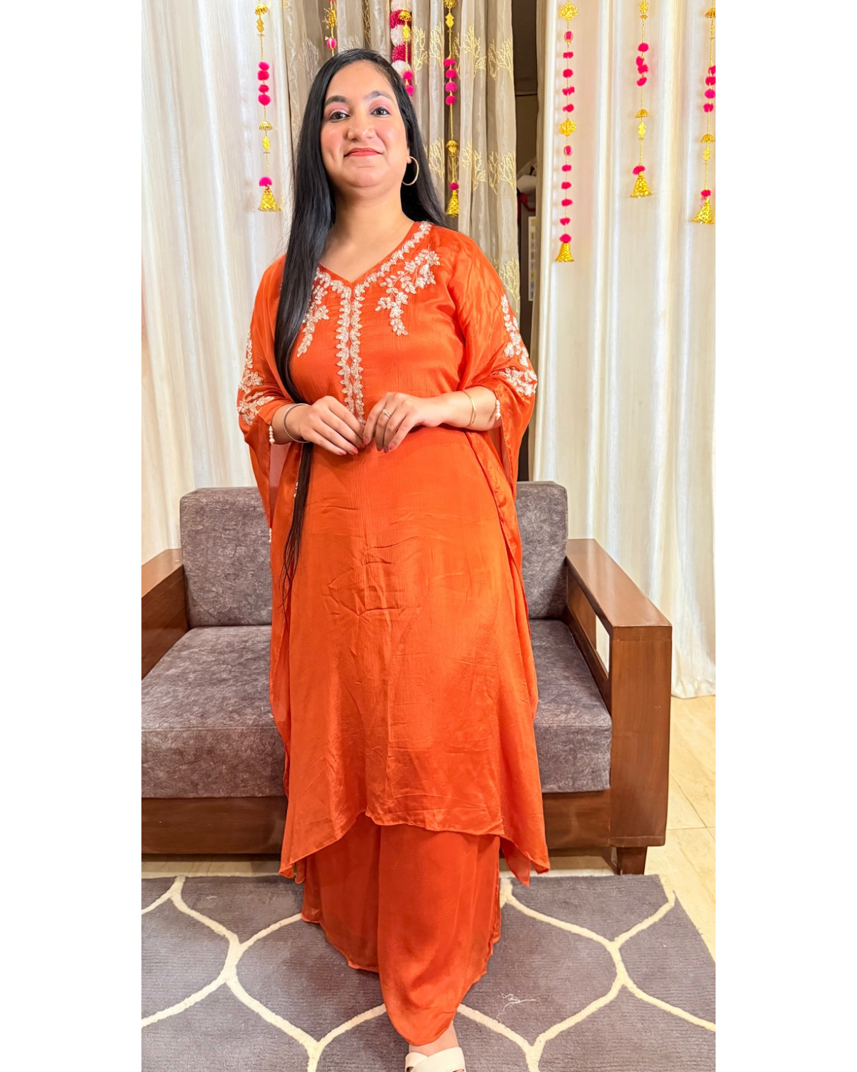 Woman in an orange traditional outfit standing in a room with a sofa and curtains. saffron shine festive kaftan set