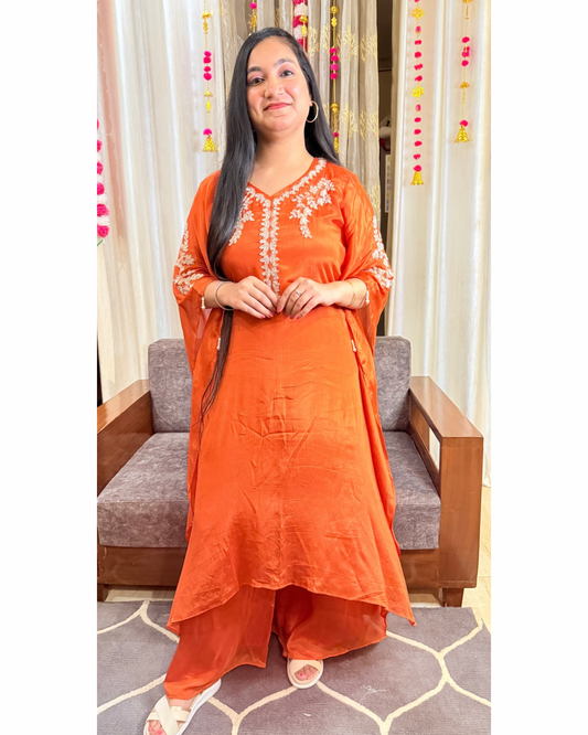 Woman wearing an orange traditional outfit standing in a room with a sofa and decorative curtains. saffron shine festive kaftan set